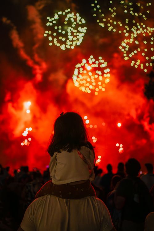 Free A child on a shoulder enjoys a mesmerizing fireworks display at night, creating a festive atmosphere. Stock Photo