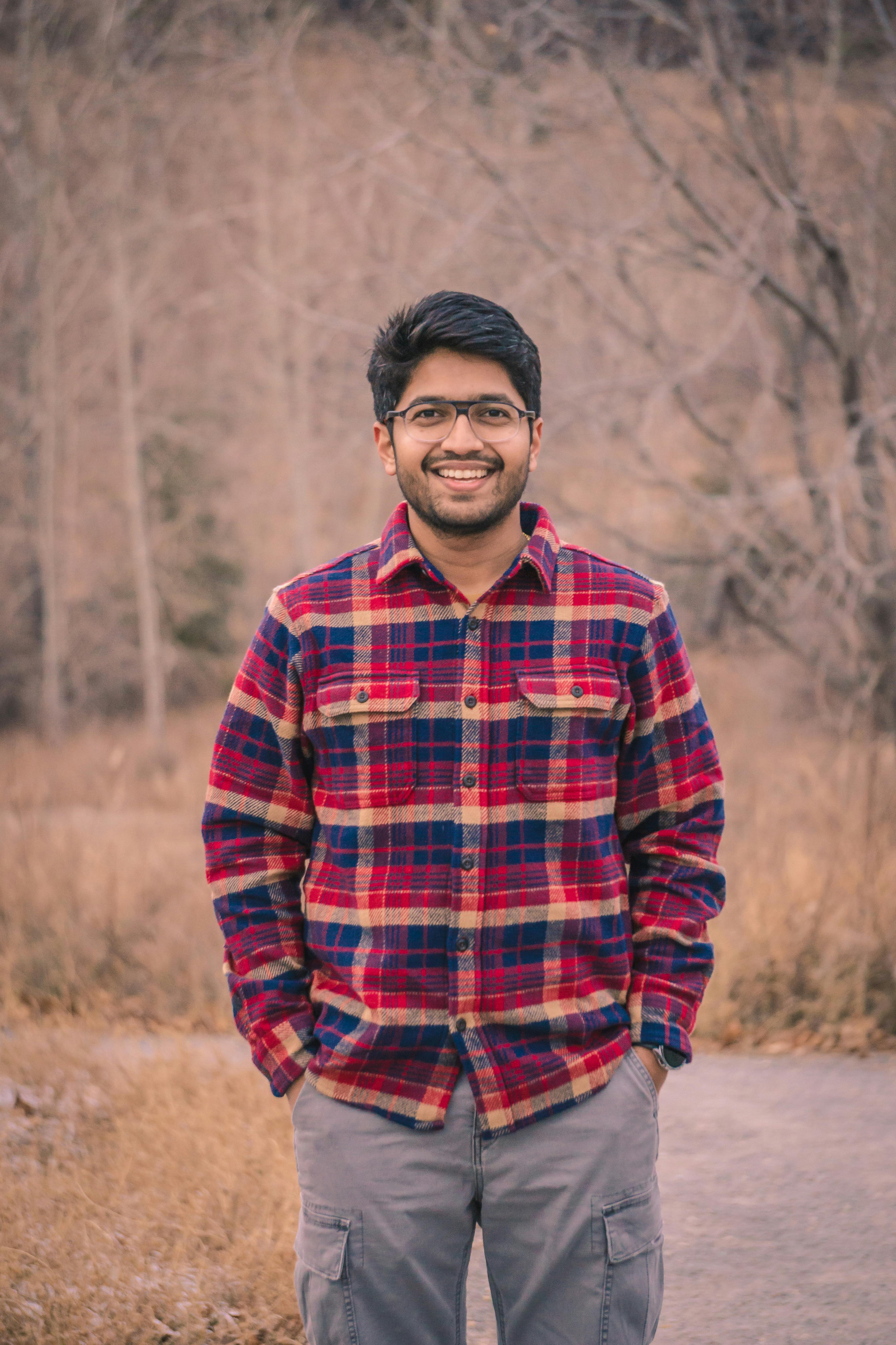 A smiling man in a red plaid shirt stands outdoors in a serene wooded area.