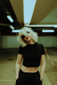 Portrait of a fashionable blonde woman wearing a black t-shirt and sunglasses, standing confidently indoors.