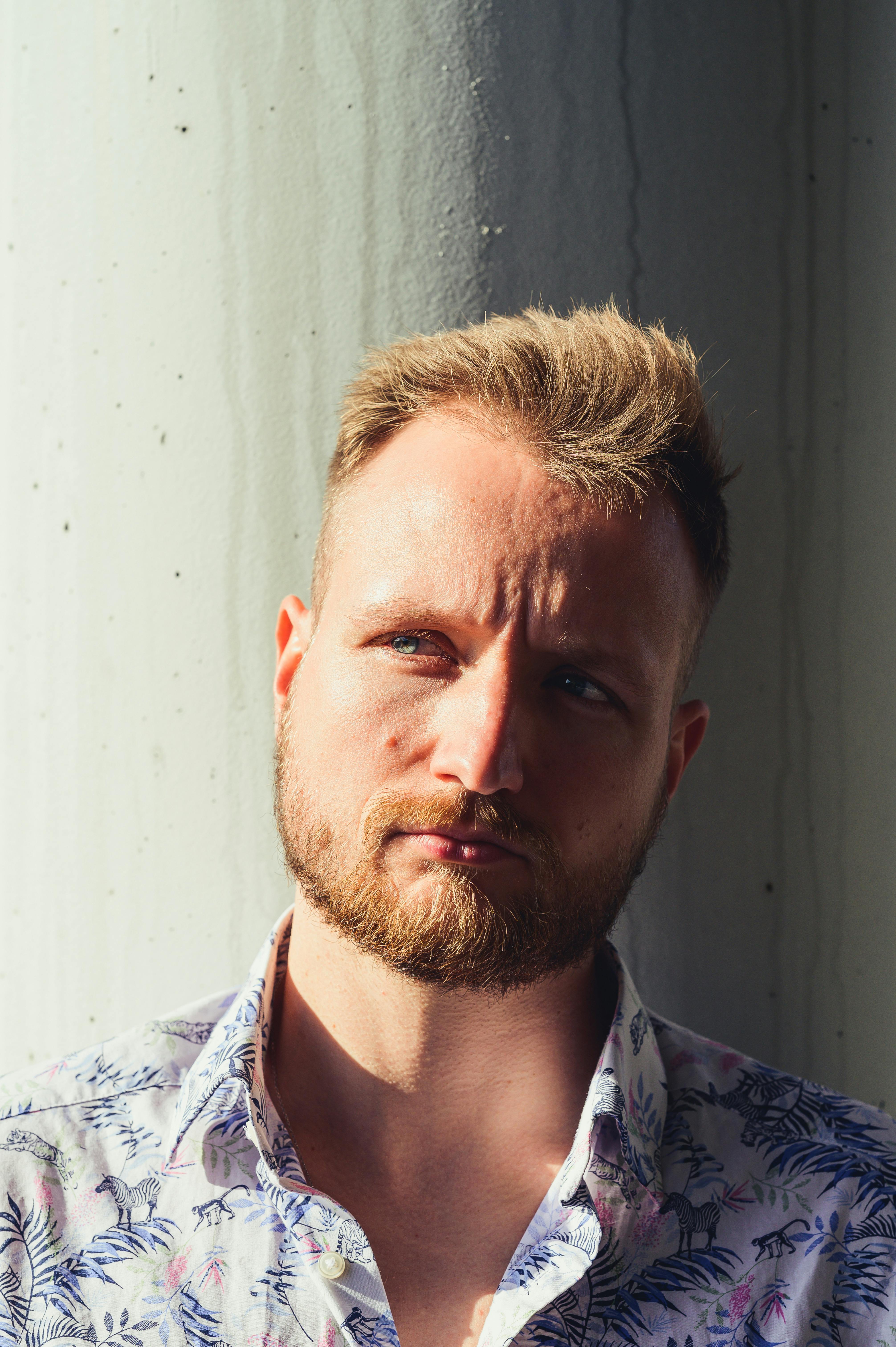 Close-up portrait of a thoughtful blonde man with a beard against a sunlit wall.