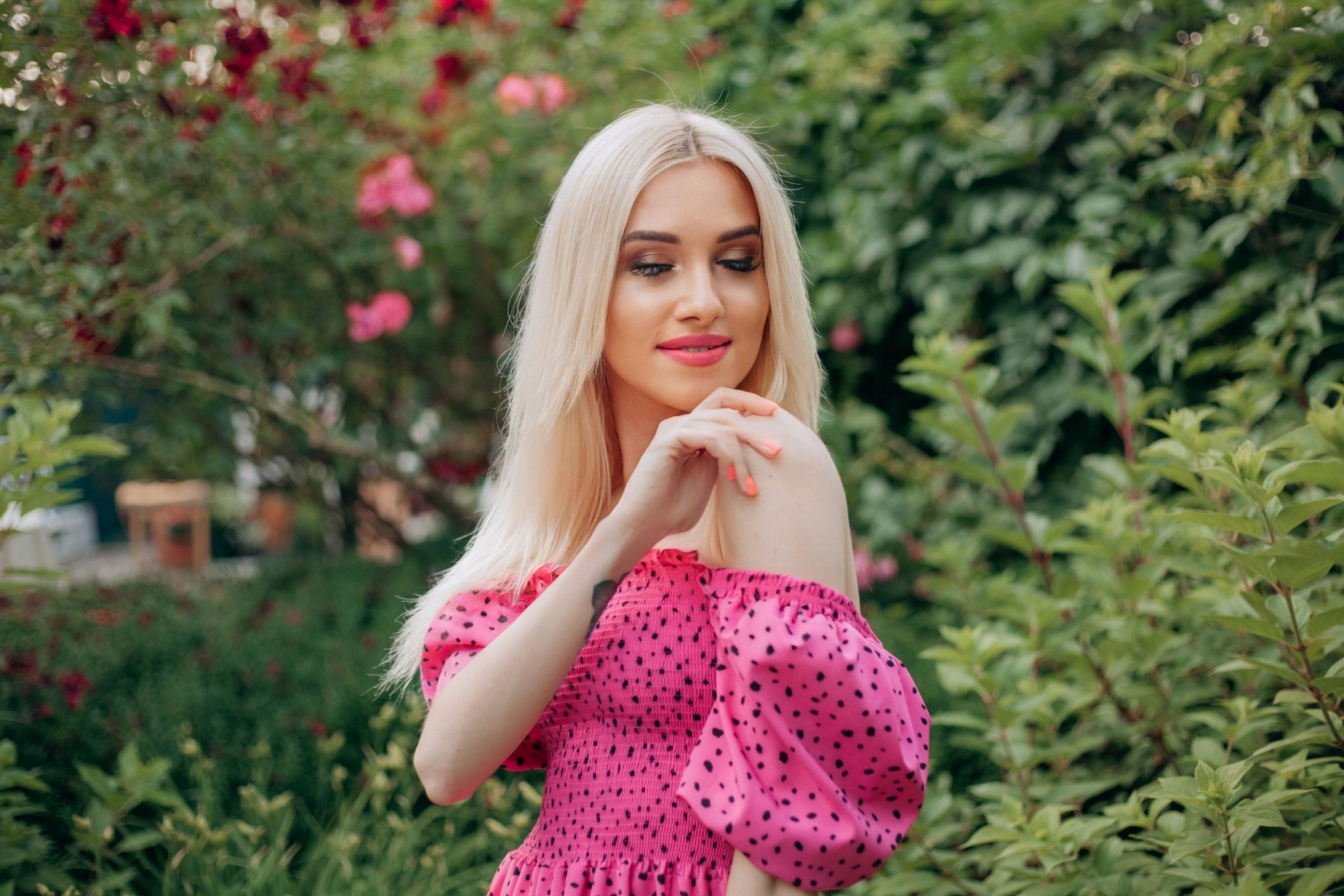 Charming portrait of a blonde woman in a pink polka dot dress standing in a lush green garden, smiling softly.