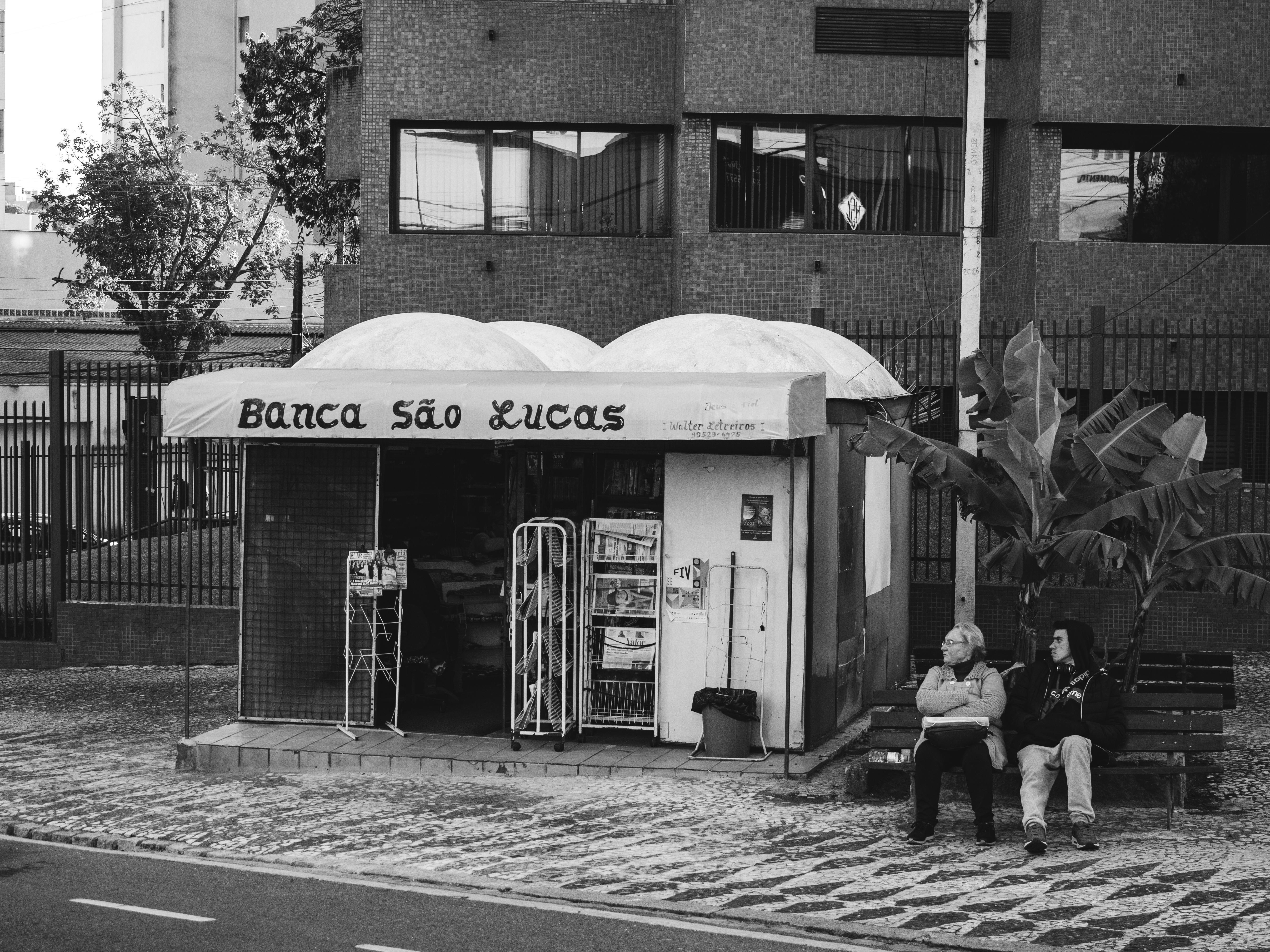 Black and white photo of a newspaper stand with people sitting on a bench.