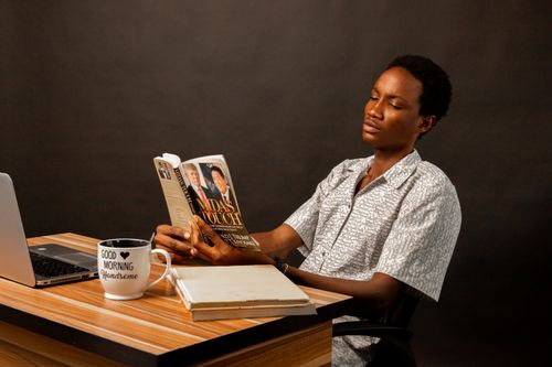 Free A young man focused on reading a book at a desk with a laptop and coffee mug, creating a studious atmosphere. Stock Photo