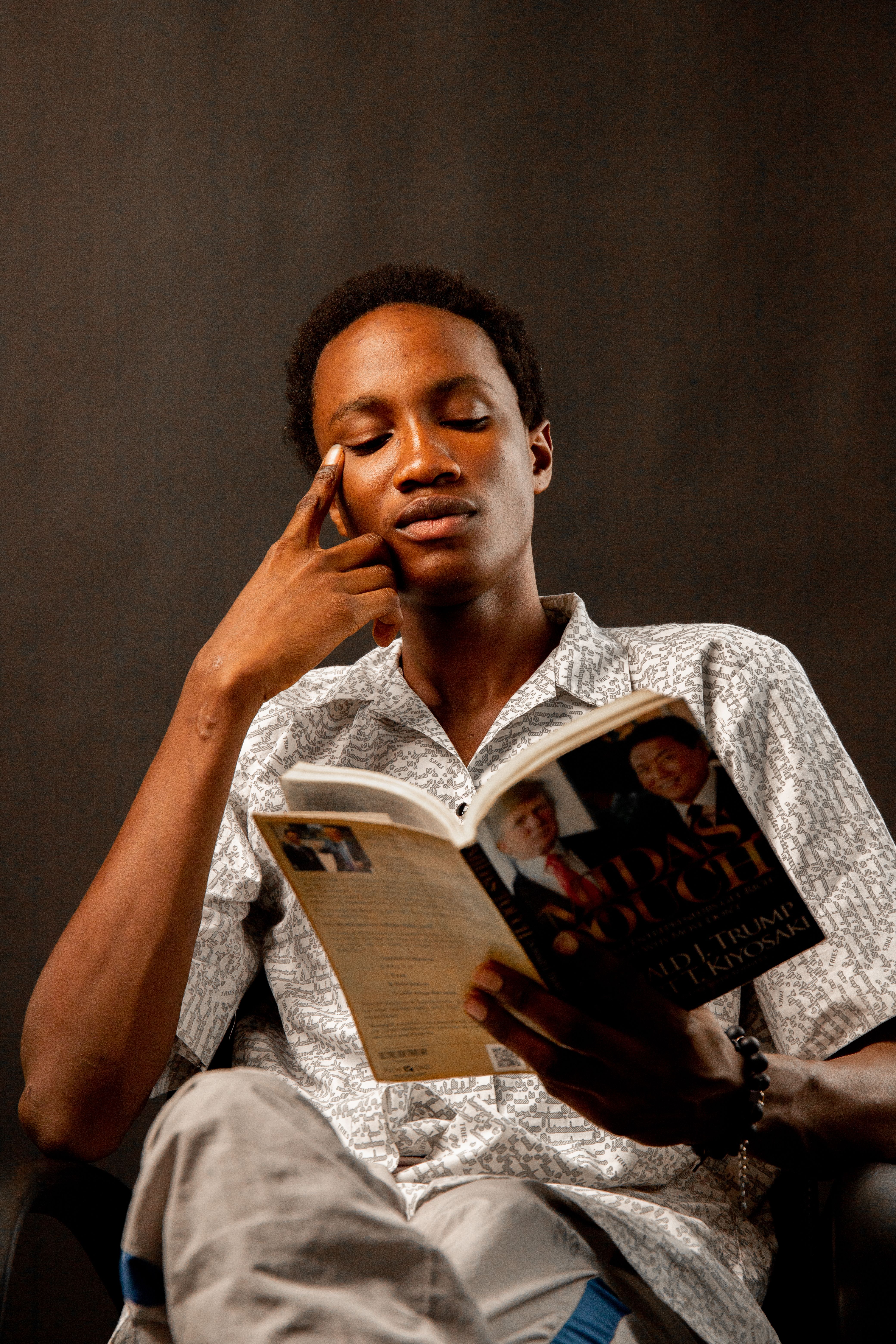 A young man sits indoors reading a book, deeply focused and relaxed in a serene setting.
