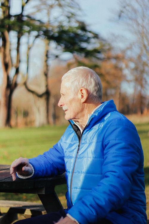 Free Senior man smiling, wearing blue jacket, sitting in a sunlit autumn park. Stock Photo