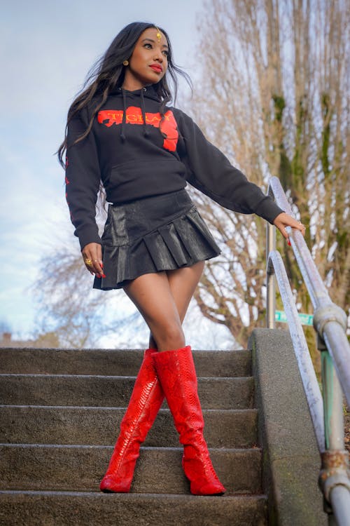 Young Model in a Black Hoodie Mini Skirt and Red Knee-high Heeled Boots on the Stairs