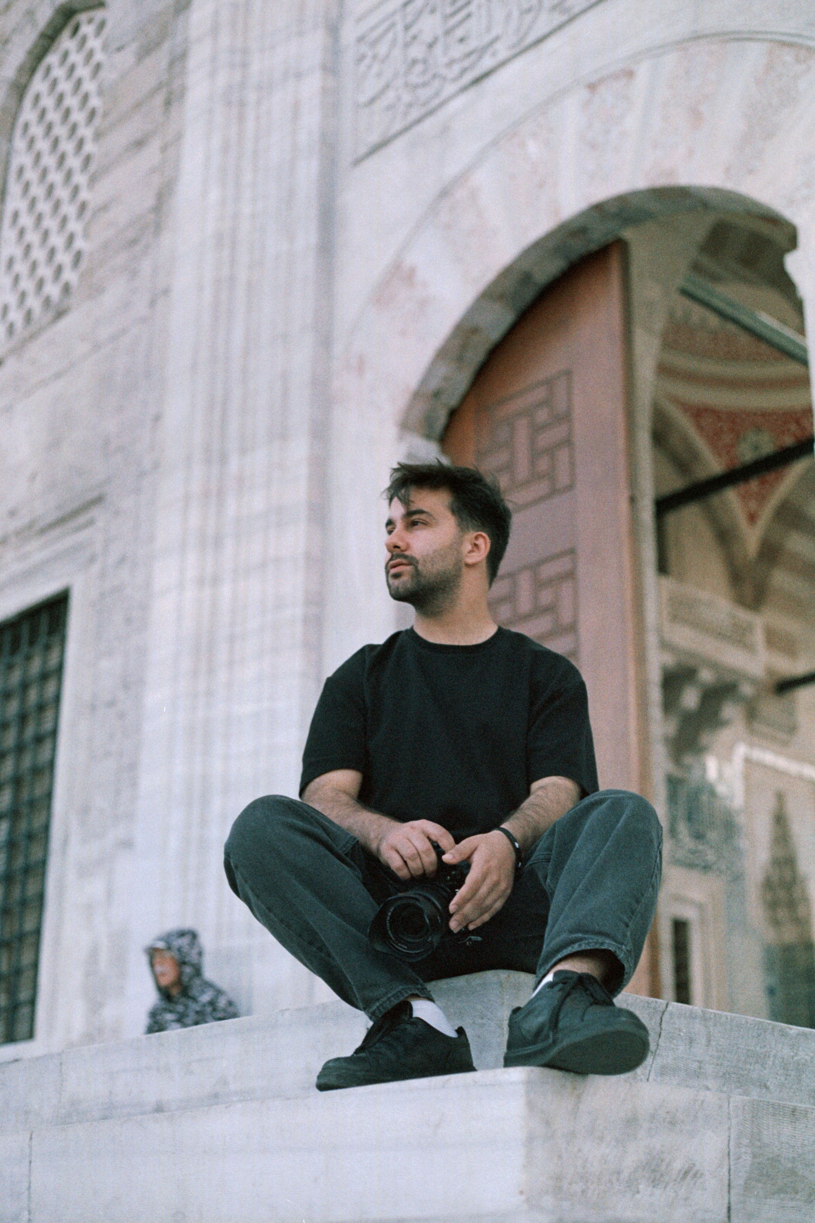 A man in a black t-shirt sits on steps outside a mosque entrance, urban setting.