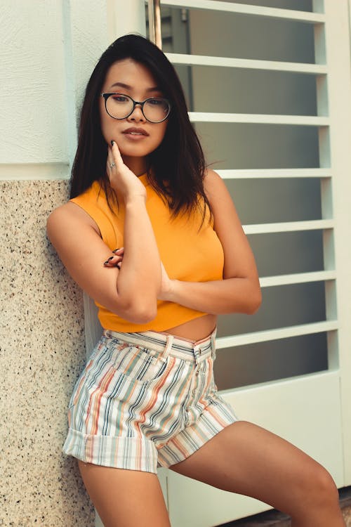 Woman in Eyeglasses, Orange Sleeveless Top, and Striped Shorts Near Window