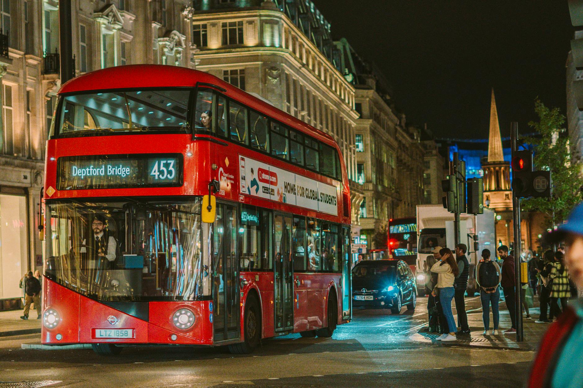 A classic red double-decker bus navigates through London's vibrant Oxford Street at night, with bustling city life around.