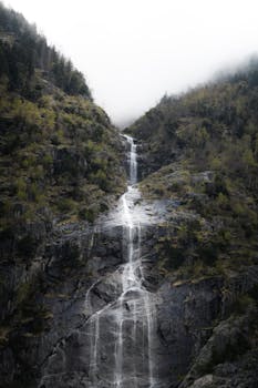 Capture the serene beauty of a misty waterfall in Ginzling, Tirol, set against lush green mountains and soothing clouds.