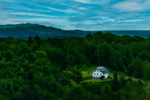 A picturesque white house surrounded by dense green forest with mountains in the background under vibrant blue skies.