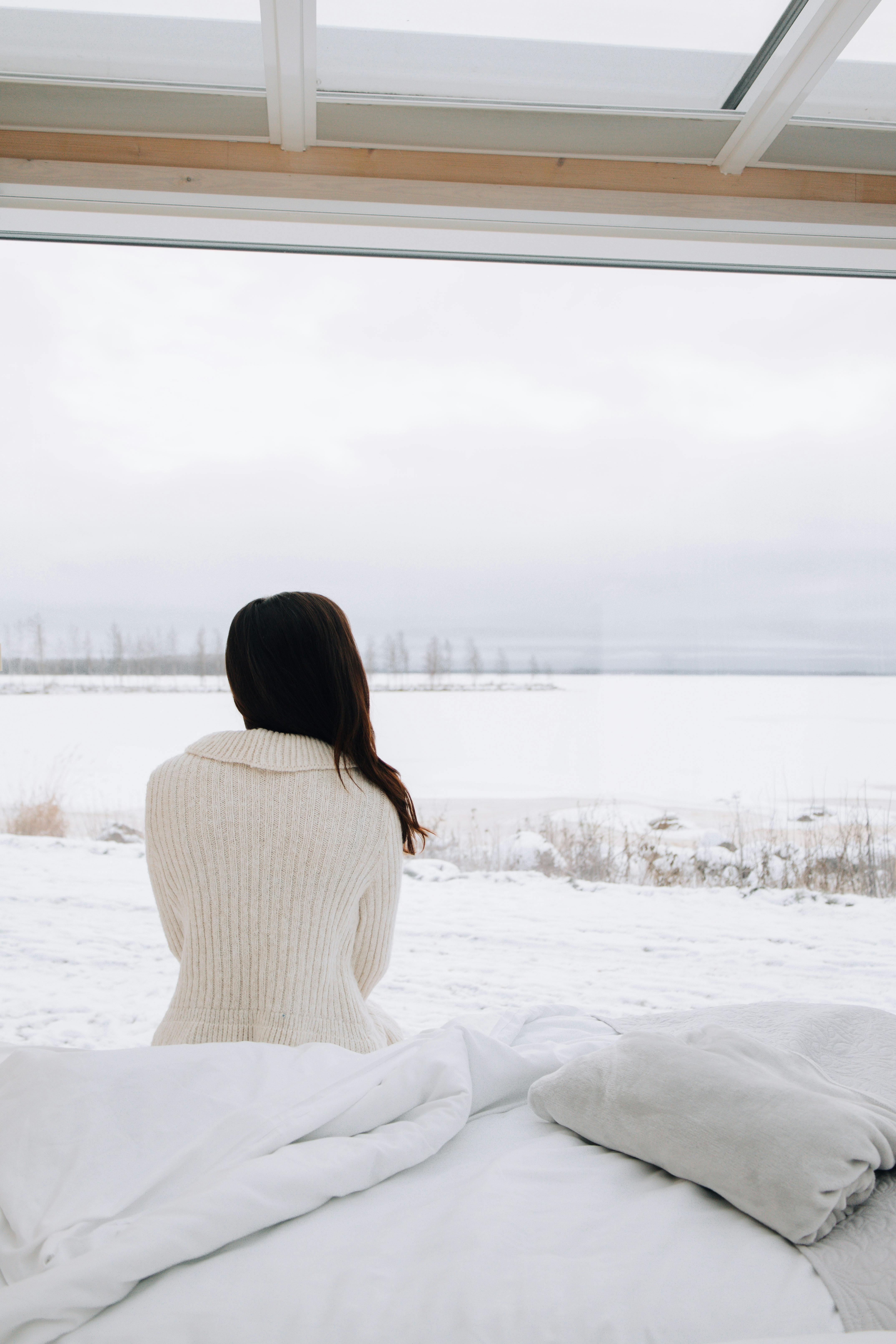 A woman in a cozy sweater sits on a bed, gazing at a snowy winter landscape in Finland.