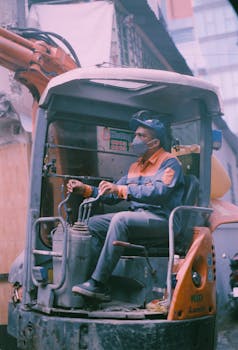 A male construction worker operates an excavator on a busy construction site, wearing a face mask and helmet.