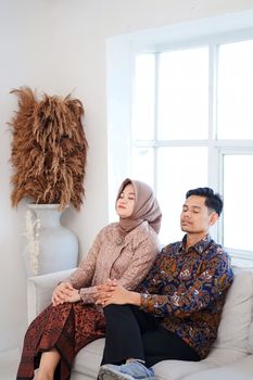 A serene Muslim couple sitting together on a couch, eyes closed, enjoying a peaceful moment indoors. - Fotografer Sidoarjo