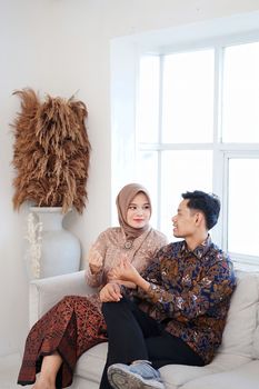 A couple in elegant traditional outfits sitting on a sofa in a bright room. - Fotografer Sidoarjo
