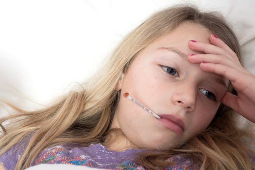 A young girl rests in bed with a thermometer, illustrating signs of illness and fatigue., Photographer: Jason Deines