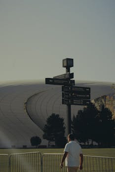 A man walks near a modern architectural landmark with a signpost in the foreground during evening light.
