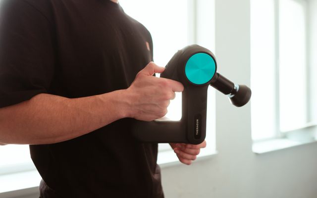 A man holding a black Theragun with a teal disc and triangular handle by a bright window