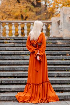 Blonde woman in a flowing orange dress standing on a stone staircase surrounded by autumn foliage.