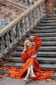 Stylish blonde woman in an orange dress posing on historic stone steps with an autumn backdrop.