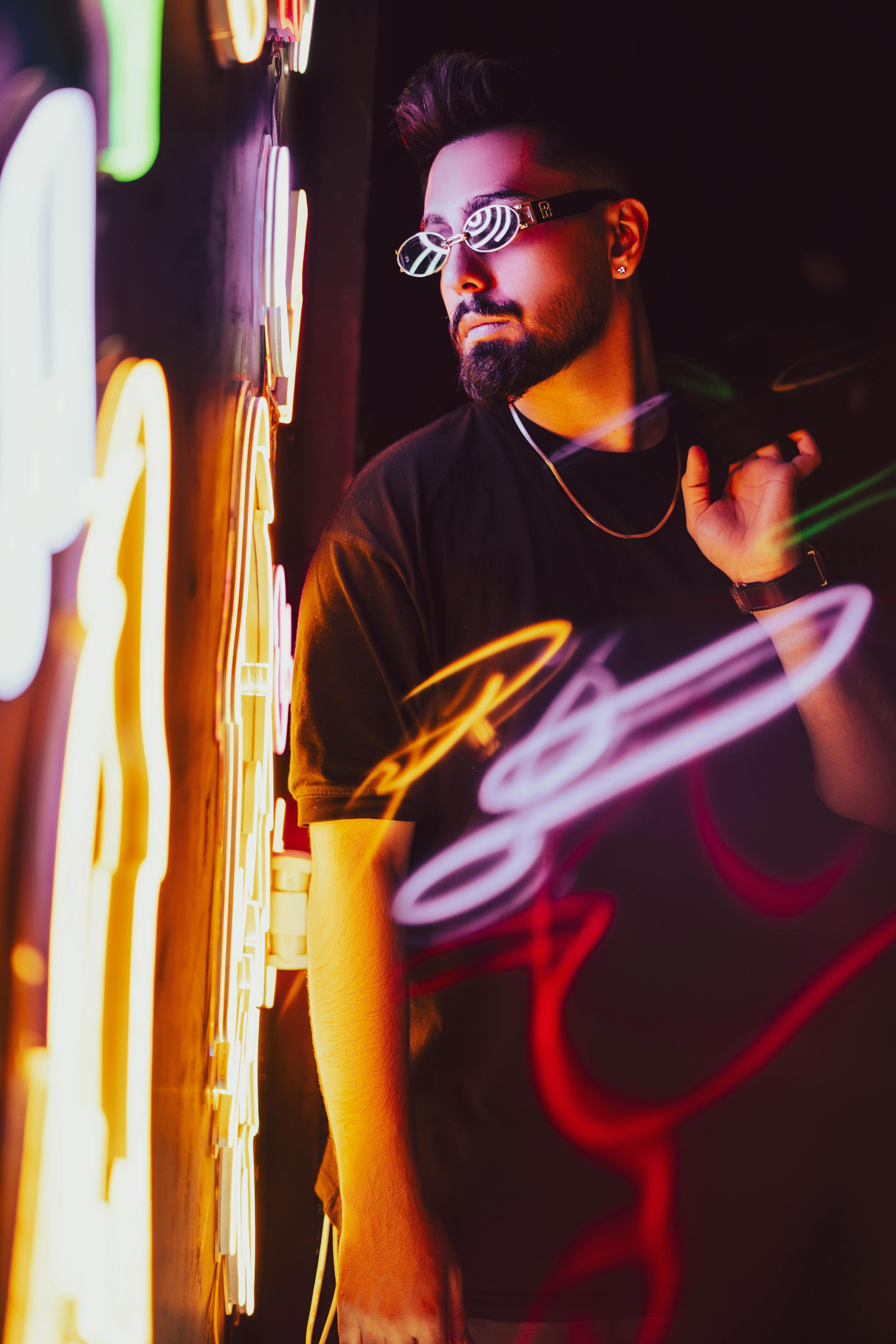 Man with sunglasses and stubble posing by neon lights in a dark setting, exuding fashion and style.