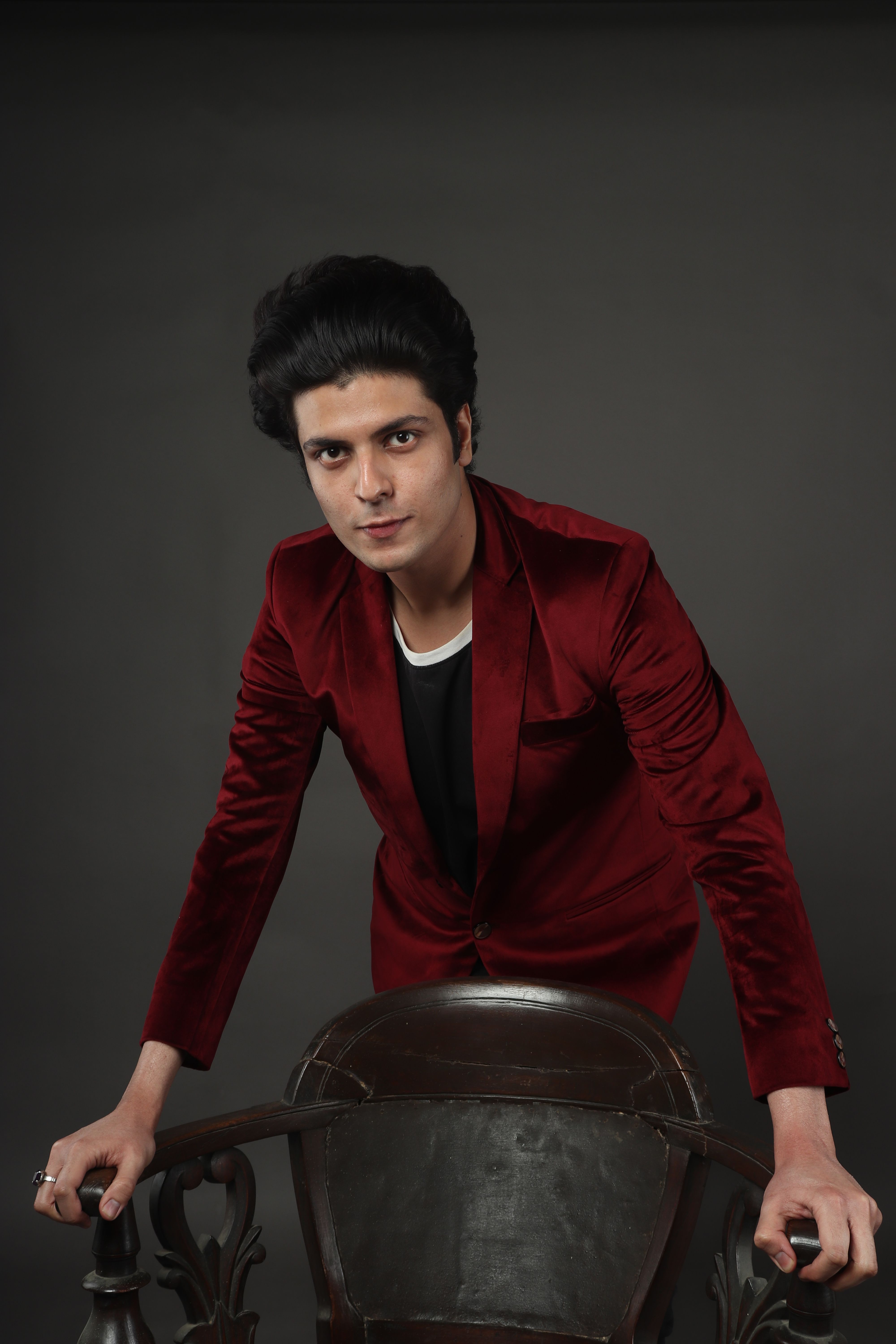 Young man in a sophisticated red suit leaning over a chair against a gray background, exuding confidence.