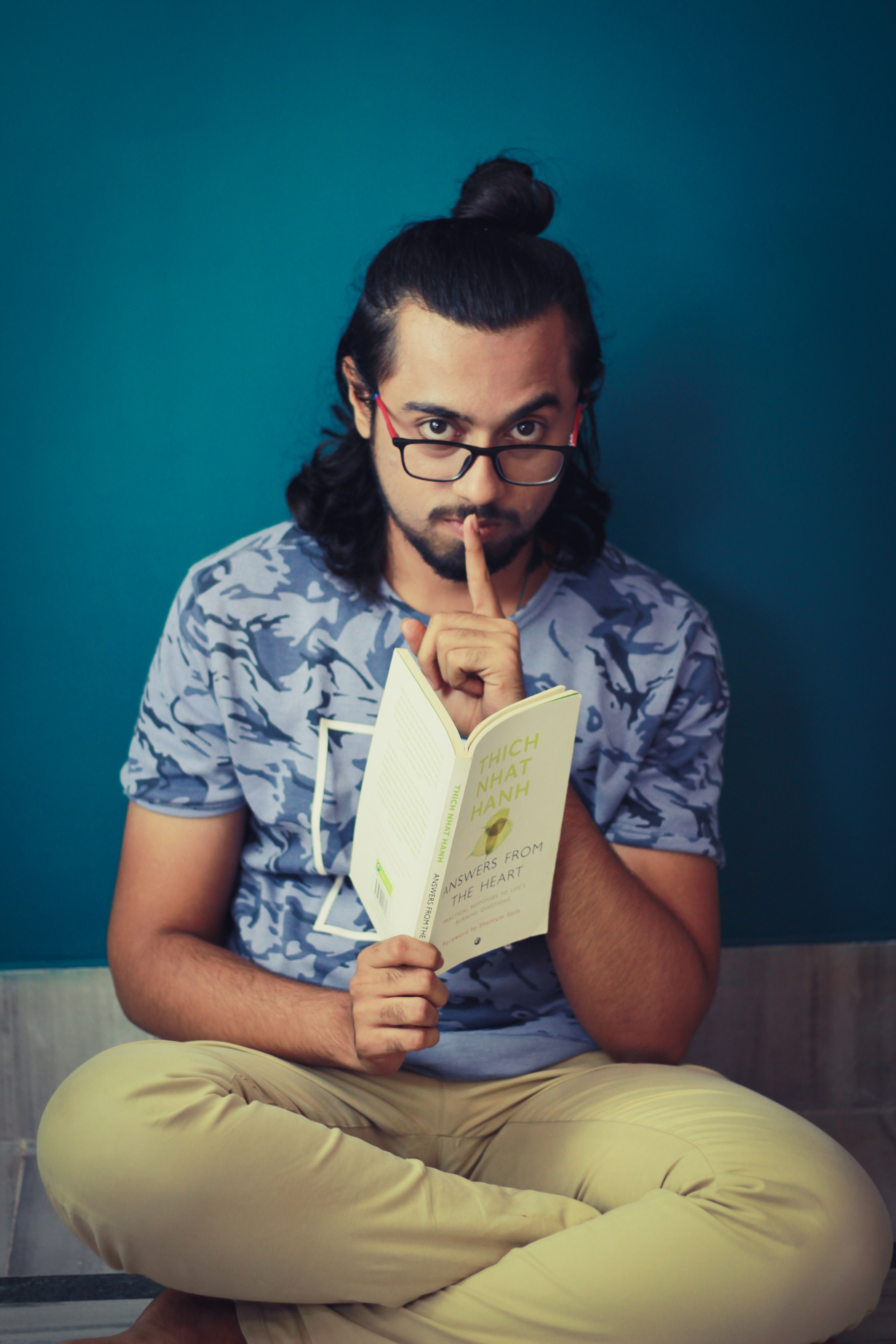 Man wearing glasses holds book, makes silencing gesture indoors against blue wall.