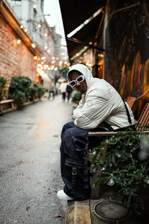 Model in Thick Frame Sunglasses Wearing a Padded White Jacket and Wide Cargo Pants Sitting in an Alley