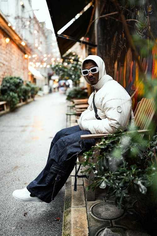 Model in Sunglasses Wearing a Padded White Jacket and Wide Cargo Pants Sitting on a Chair in an Alley