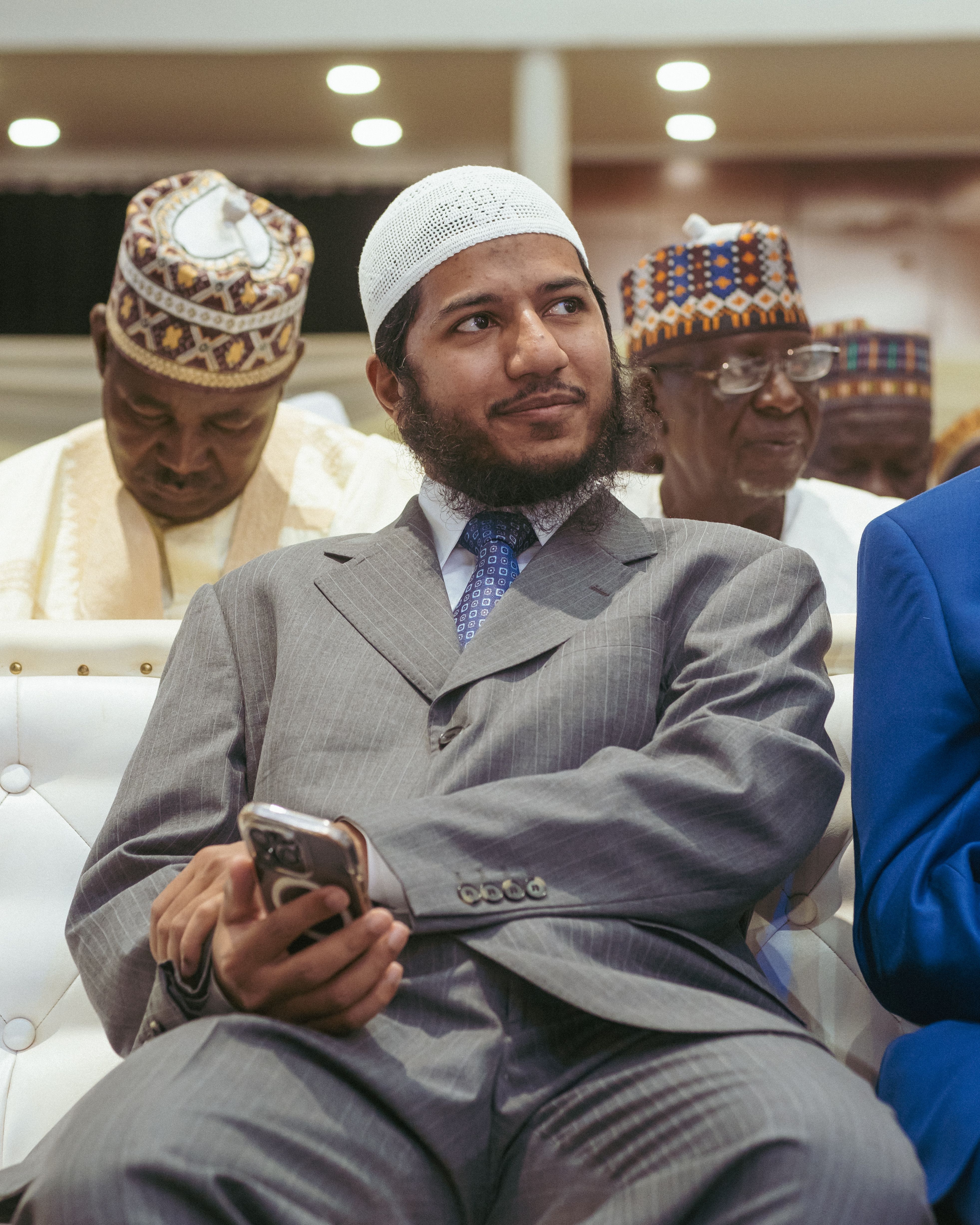 A man in a gray suit and white cap sits holding a smartphone at an indoor event.