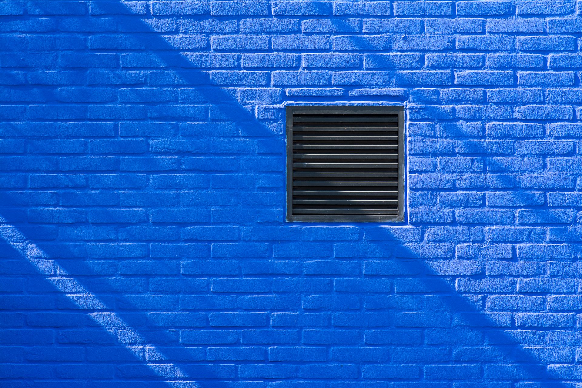 Close-up of a bright blue brick wall featuring a black ventilation grate and shadows.