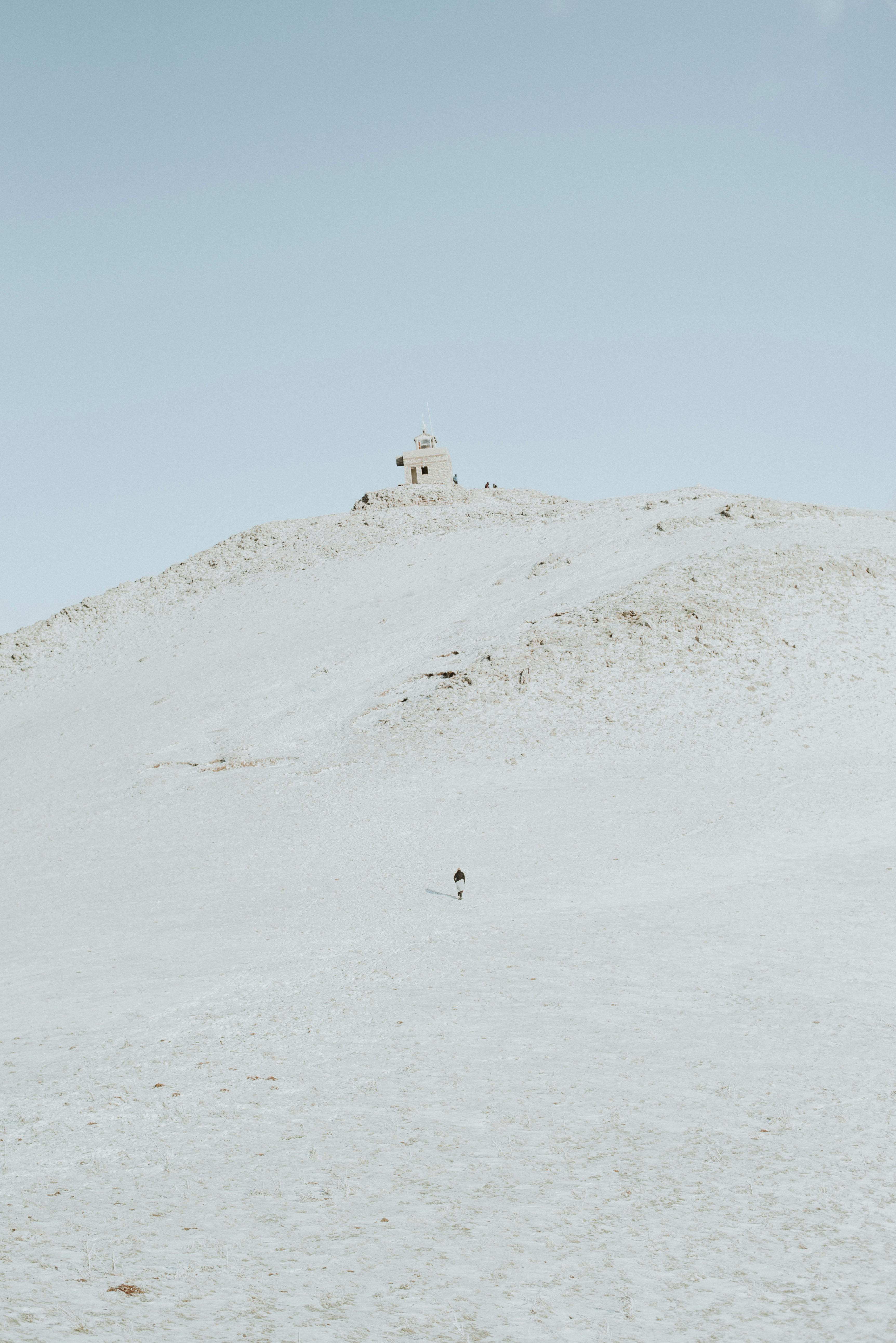 A solitary person walks up a snowy hill toward a small distant building under a clear winter sky.