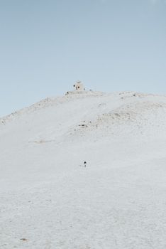 A solitary person walks up a snowy hill toward a small distant building under a clear winter sky.