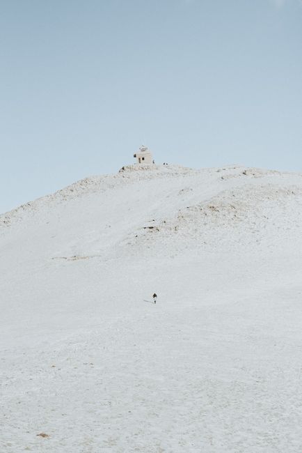 A solitary person walks up a snowy hill toward a small distant building under a clear winter sky.