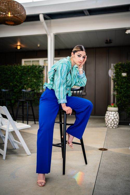 Free Fashion model posing on a stool, wearing blue pants and a striped shirt against a modern outdoor setting. Stock Photo