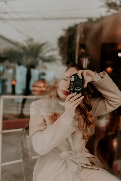 Free A woman in a coat takes a photo through a glass window, blending urban and artistic vibes. Stock Photo