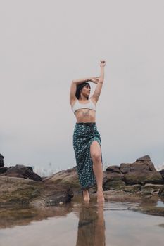 A woman in a summer outfit poses confidently on rocky terrain by the water.