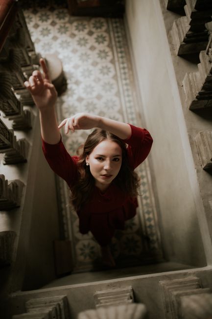 Portrait of a woman in a red dress looking up while standing on an ornate staircase.