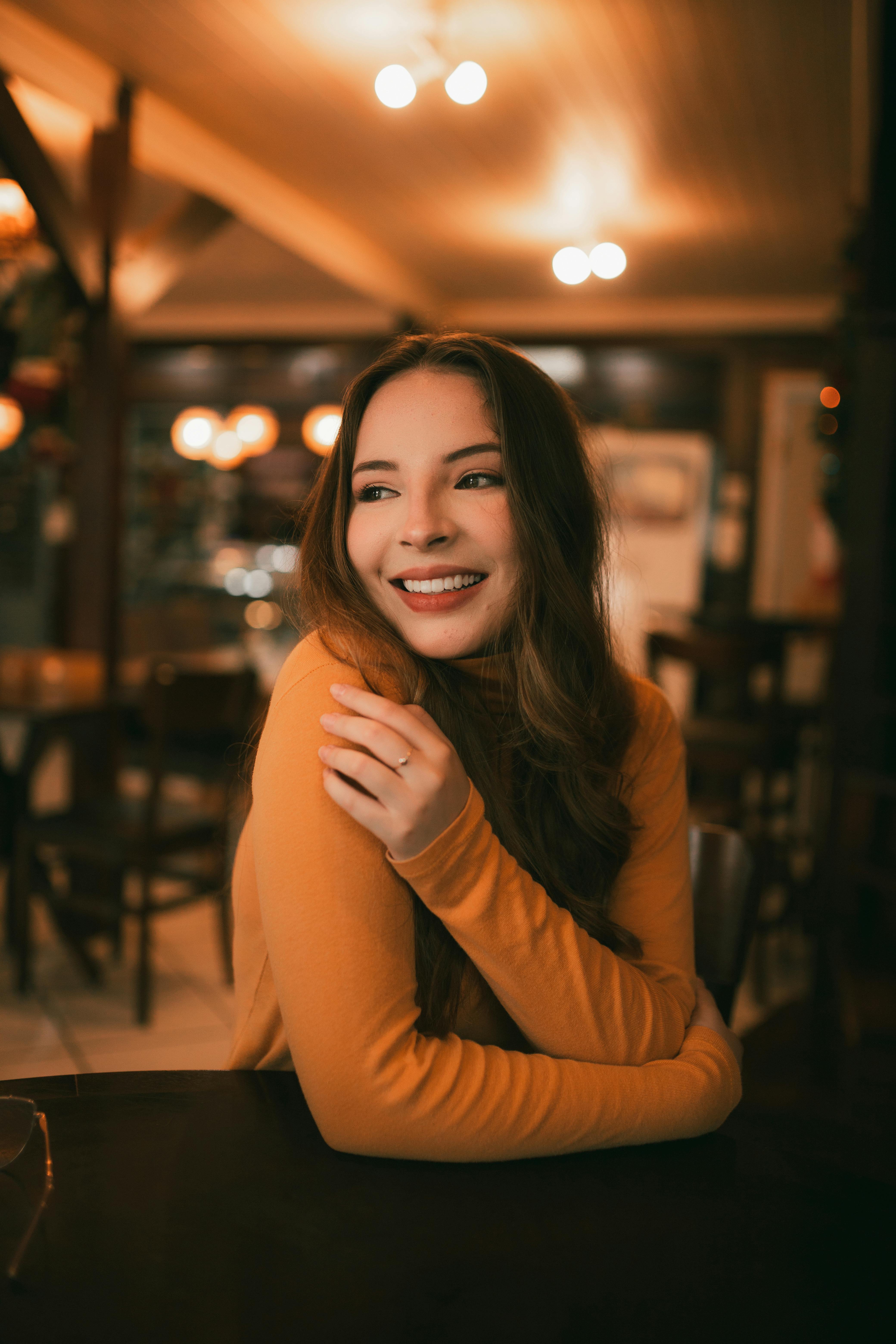 A woman in an orange sweater smiles warmly in a cozy, dimly lit café setting.