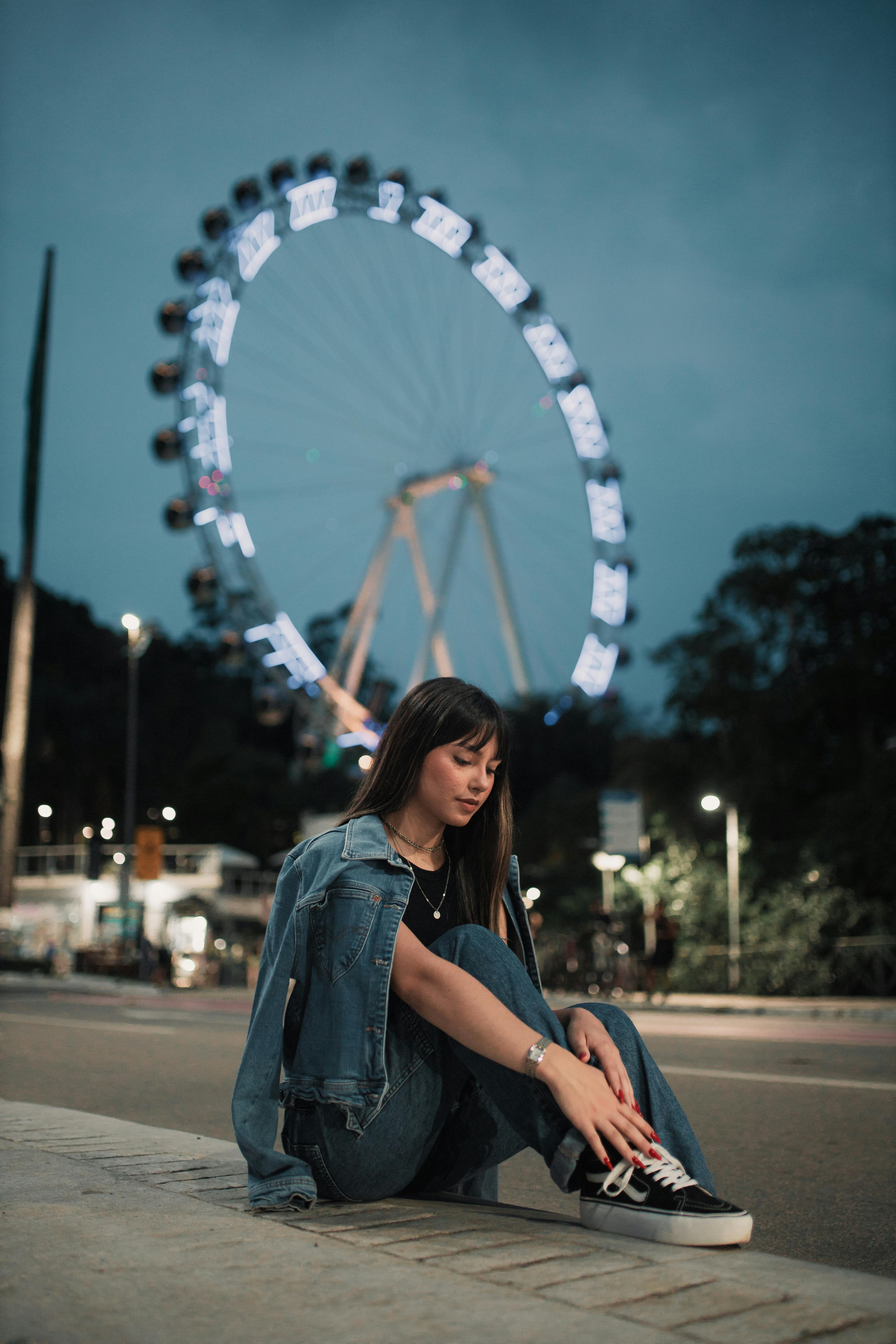 Young woman sitting near a brightly lit Ferris wheel during evening on city streets.