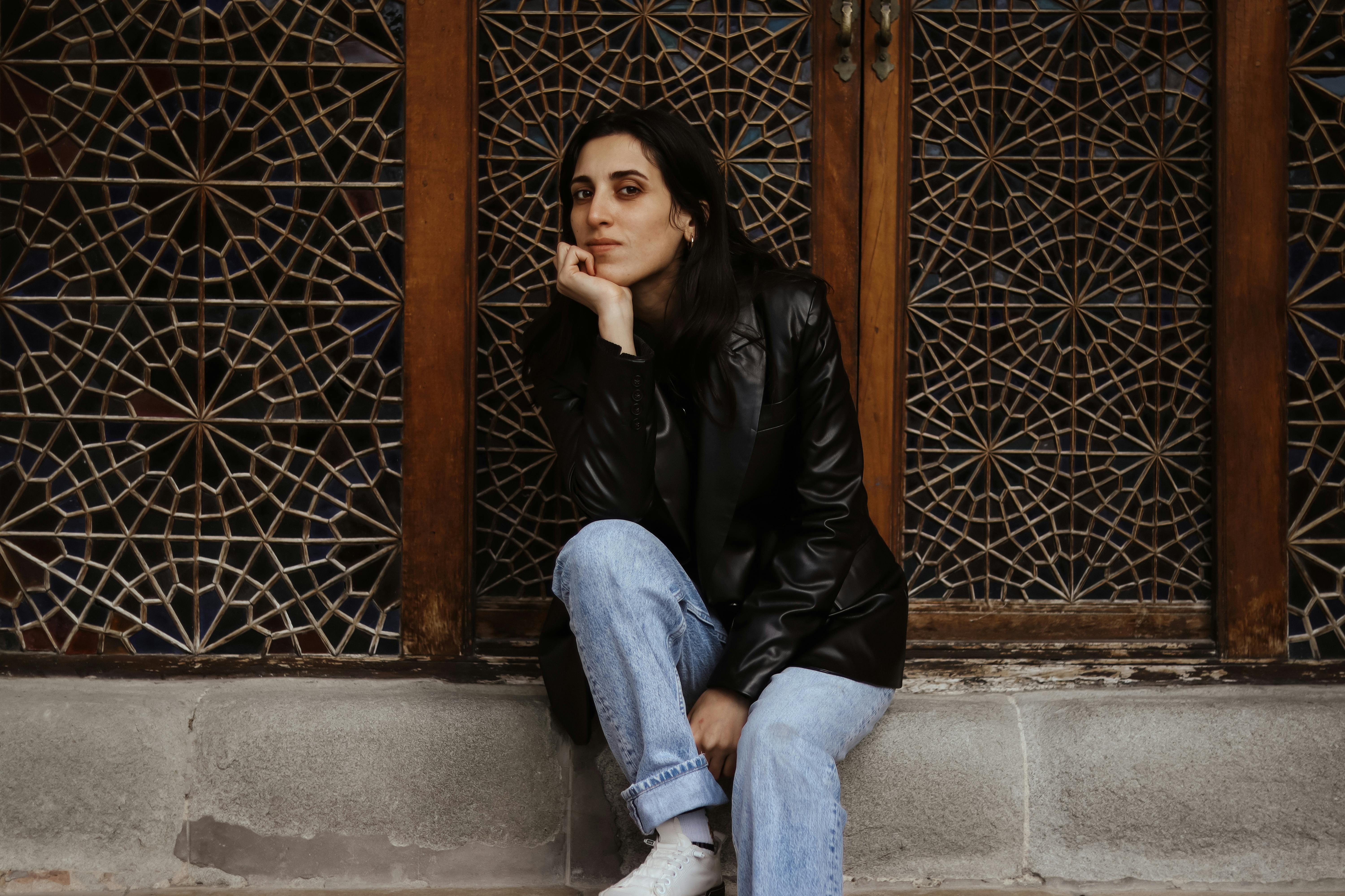 Young woman in leather jacket and jeans sitting by ornate wooden door.