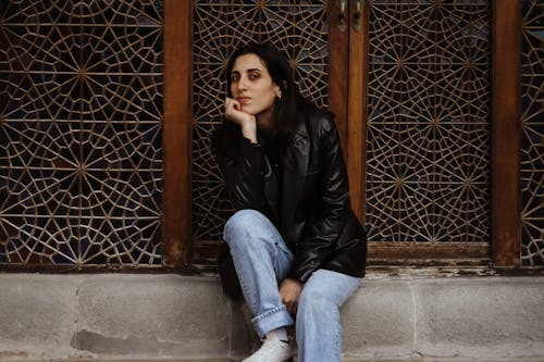 Brunette Woman in Leather Jacket and Jeans Sitting by Wall