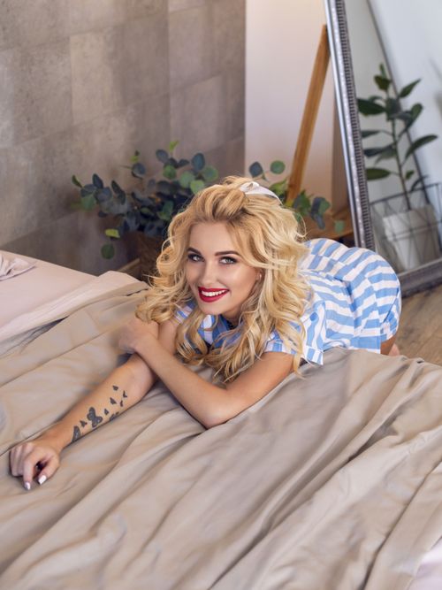 Free Smiling blonde woman in striped dress relaxing on a bed, showing tattoos, indoors. Stock Photo