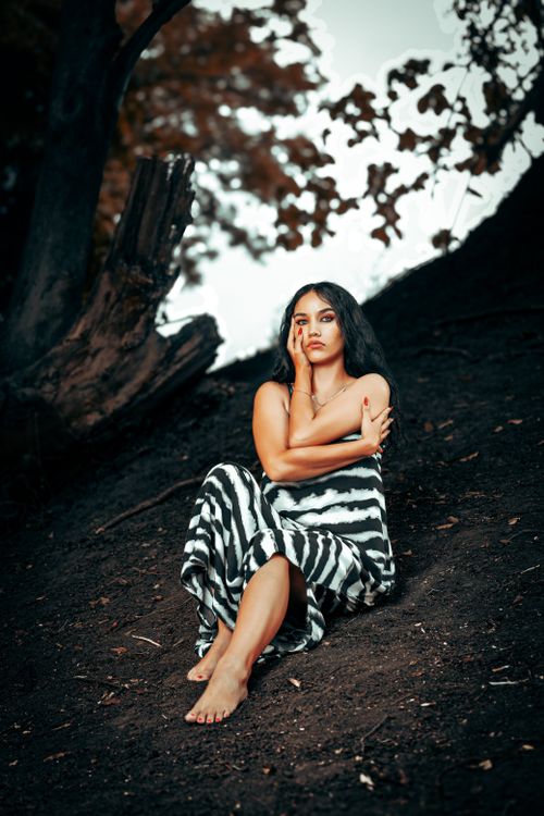 Free A woman sits barefoot in a forest wearing a zebra-striped dress, embodying fashion and nature. Stock Photo