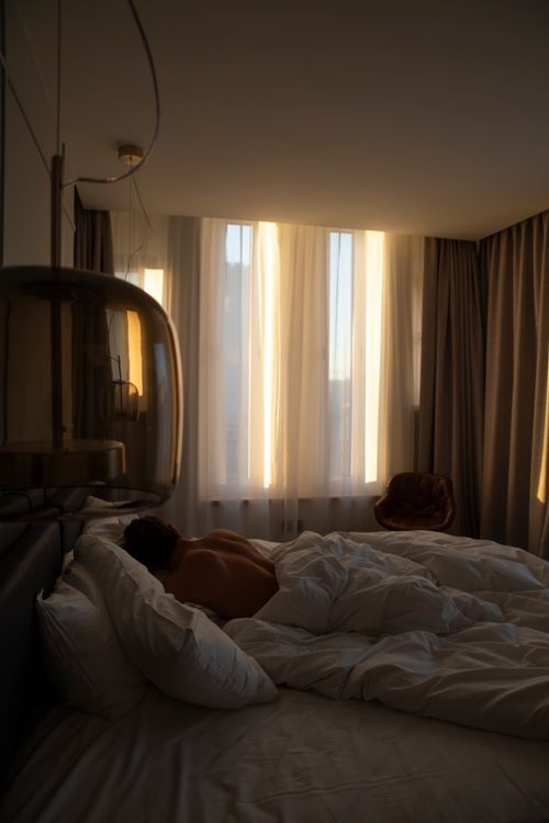 Free A shirtless man sleeps in a sunlit bedroom with soft bedding and curtains. Stock Photo