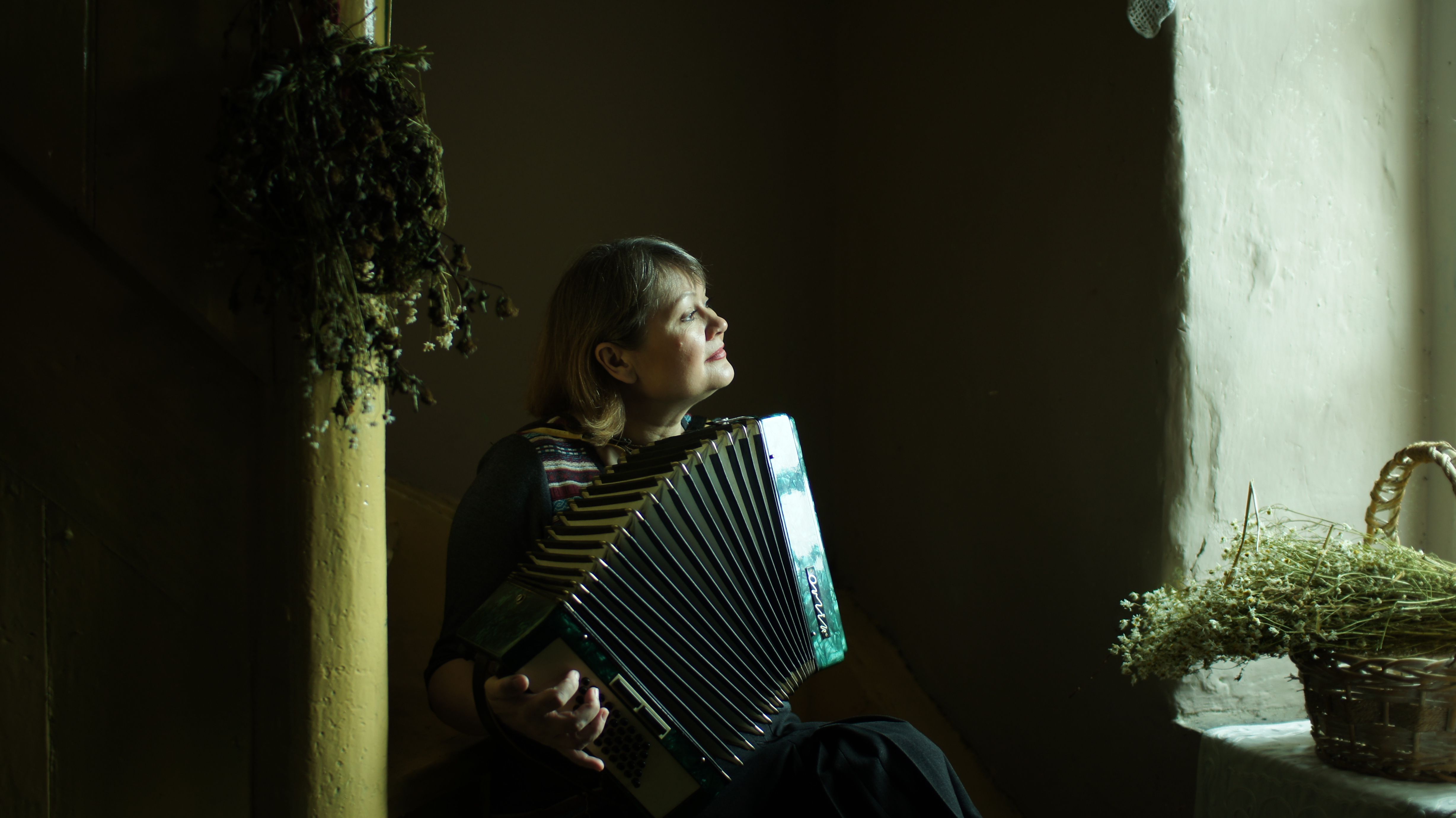 A woman sits playing an accordion, gazing thoughtfully in a dimly lit room.