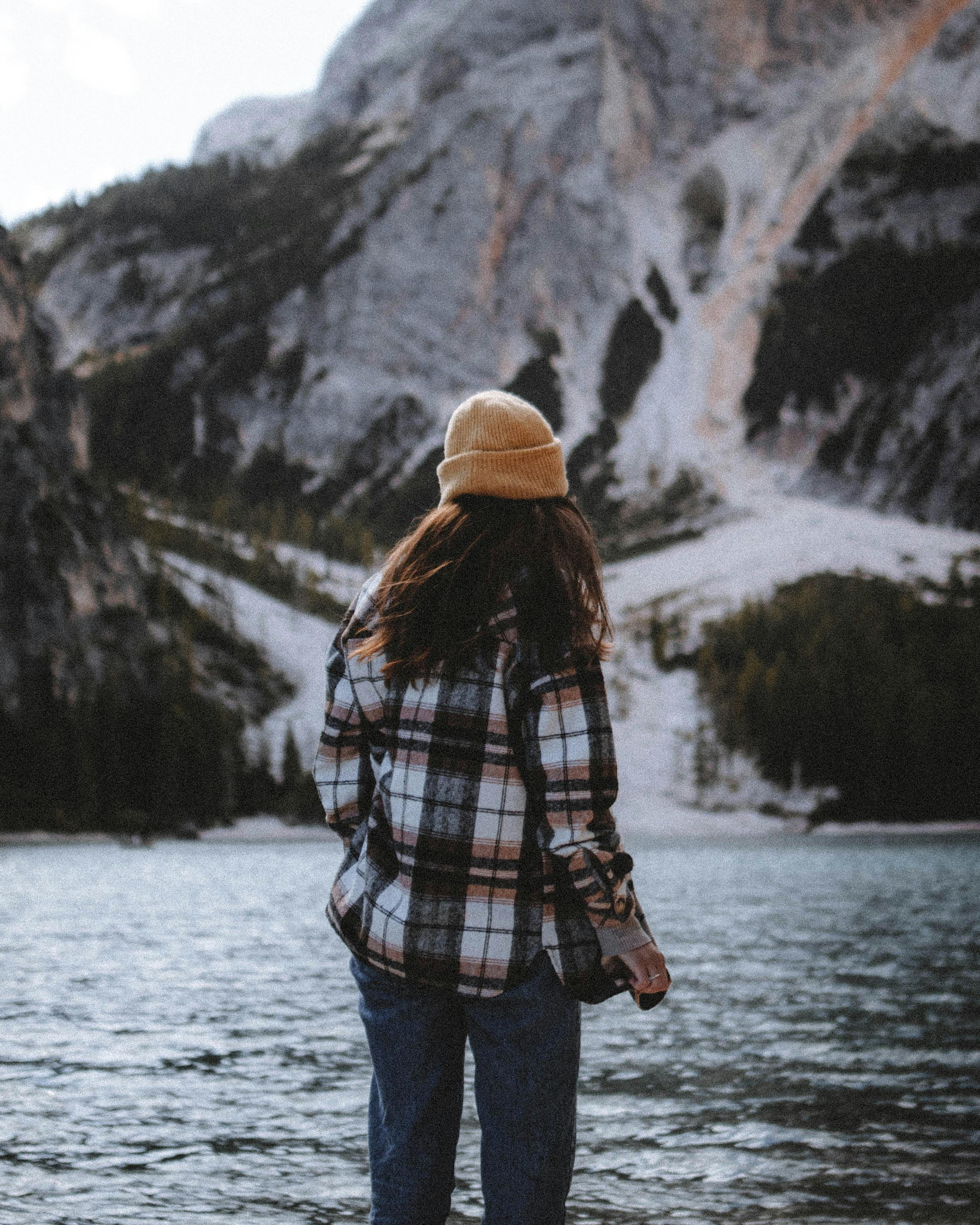 A woman in a yellow hat stands by a mountain lake, embracing the serene landscape.