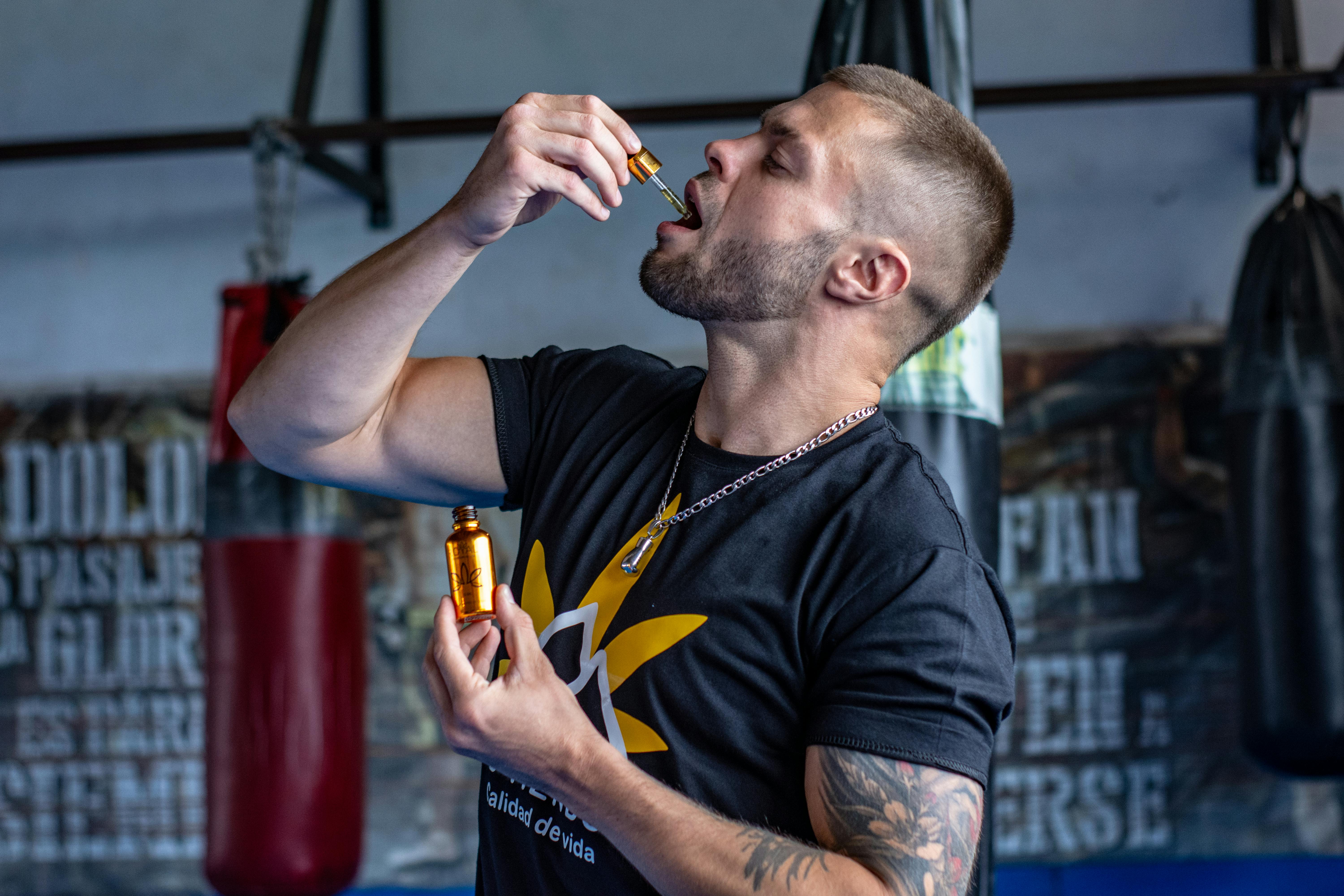 A muscular man in a gym takes a liquid supplement with a dropper.