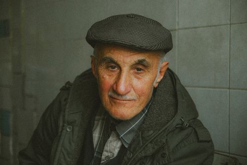 Free Elderly man with mustache and flat cap sitting indoors, looking warmly at the camera. Stock Photo