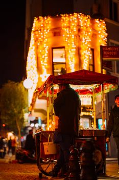 Man at a street food stand in İstanbul, Türkiye, illuminated by vibrant city lights at night.